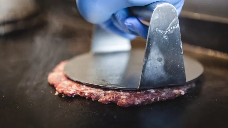 Foto de ejemplo: una mano con guante aprieta una espátula sobre la carne en la plancha